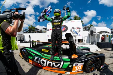 The Forbush Performance driver raises a checkered flag while standing on the No. 18 Duqueine at Road Atlanta.